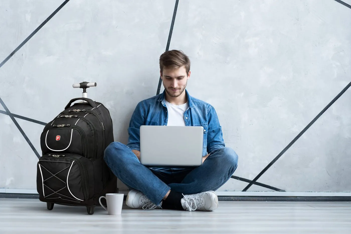 Man on his laptop sitting beside a SwissGear rolling backpack