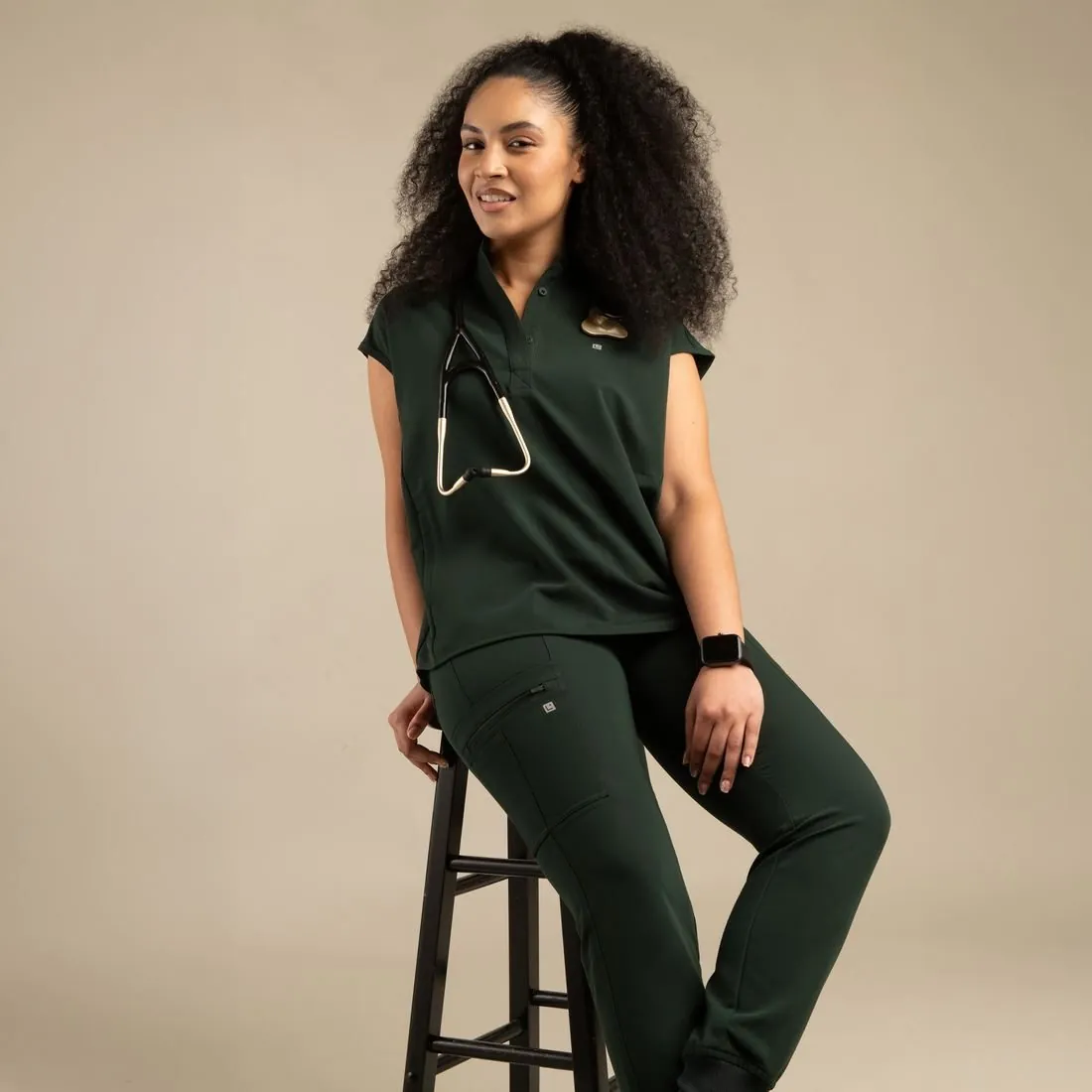 Model seated on a stool in a deep green scrub set
