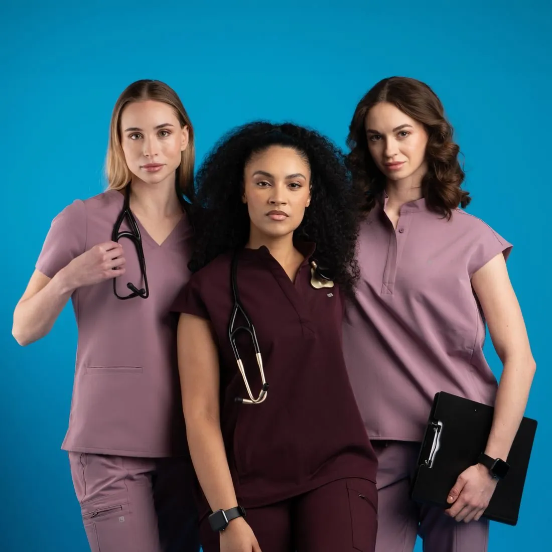 Three models in mauve and plum scrub sets against a blue backdrop